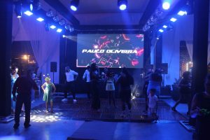 Convidados dancando na pista sob luzes azuis, globos espelhados e telao de LED