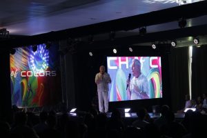 Palestrante no palco diante de plateia, com telao de LED e grade de iluminacao da Angelo Eventos